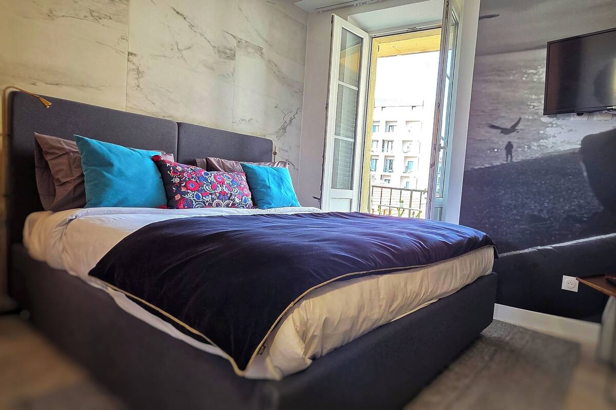 A modern bedroom with a large bed, a TV mounted on the wall, and a window with a view of buildings.