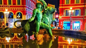 Vista nocturna de una plaza urbana con una estatua de un hombre y un caballo, rodeada de edificios con letreros iluminados.