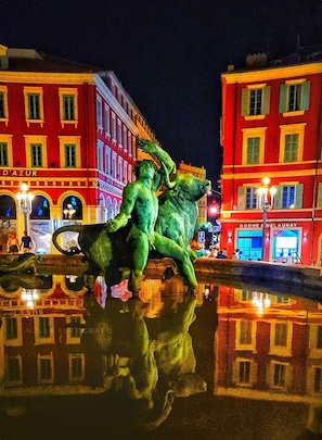 Two figures in a dynamic pose stand in a public square, surrounded by illuminated buildings and a reflective pool.