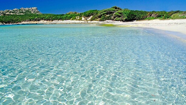 Mar turquesa y transparente con una playa de arena y afloramientos rocosos al fondo.