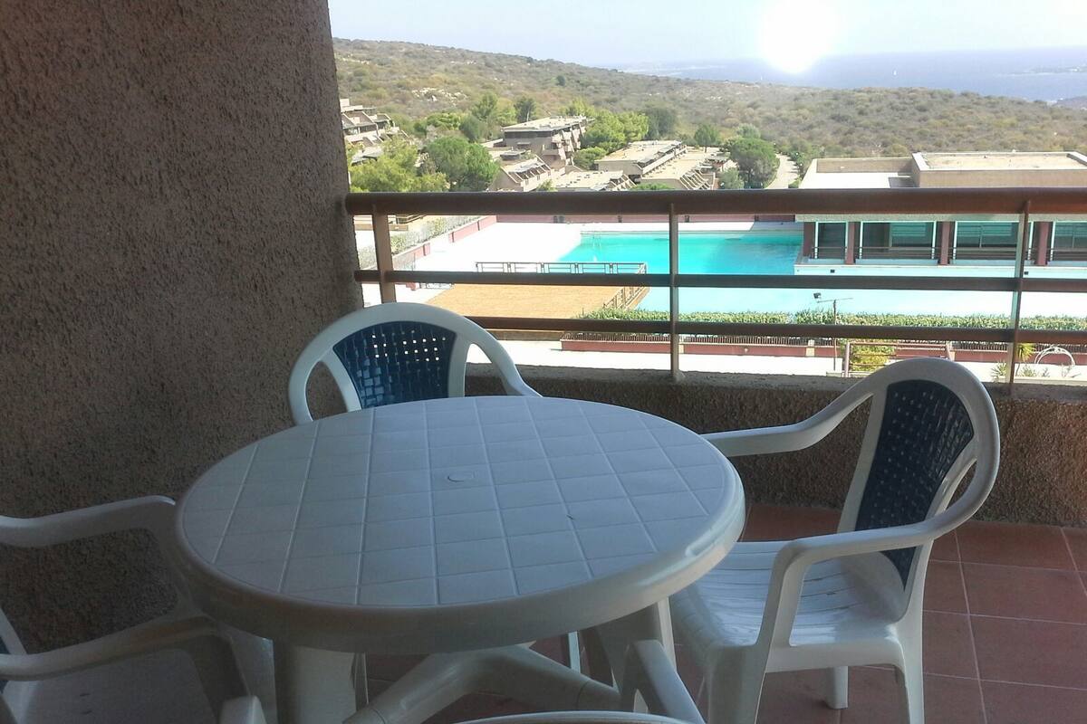 A balcony with a round table and four chairs, overlooking a swimming pool and buildings.