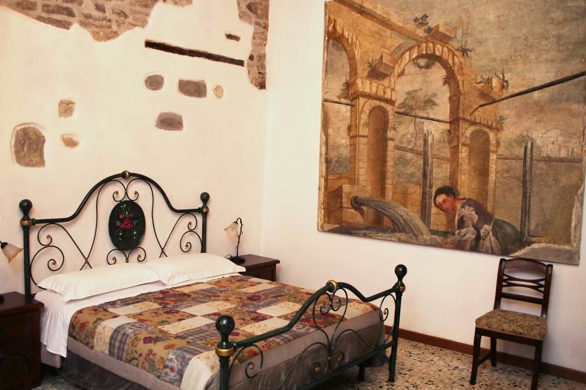 A bedroom with a bed, a chair, a nightstand, and a wall painting.