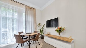 Un área de comedor moderna con una mesa redonda, dos sillas, una planta en maceta, un televisor montado en la pared y una consola de madera con un jarrón de flores.