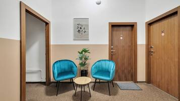 Quarto com duas cadeiras azuis, uma mesinha, um vaso de planta e duas portas de madeira com números.