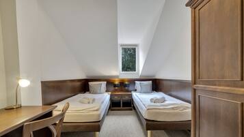 A bedroom with two single beds, a desk, and a chair. There is a window providing natural light and a small table with two lamps.