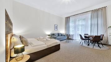 A modern hotel room with a bed, a sofa, a dining table with chairs, and a large window with curtains.