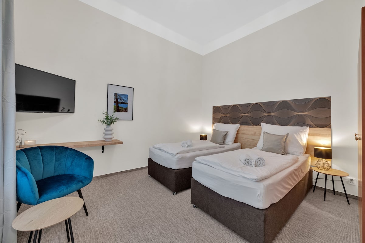 A hotel room with two beds, a TV, a blue chair, a small table, and a wall-mounted shelf.