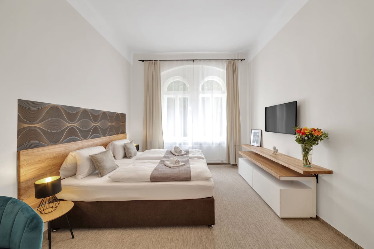A modern hotel room with a large bed, a bedside table with a lamp, a TV mounted on the wall, and a small desk with a vase of flowers.