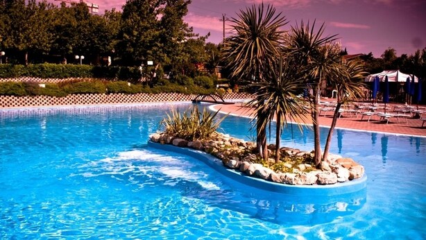 A swimming pool with a small island featuring palm trees and rocks. - Garden Village San Marino (Borgo Maggiore)