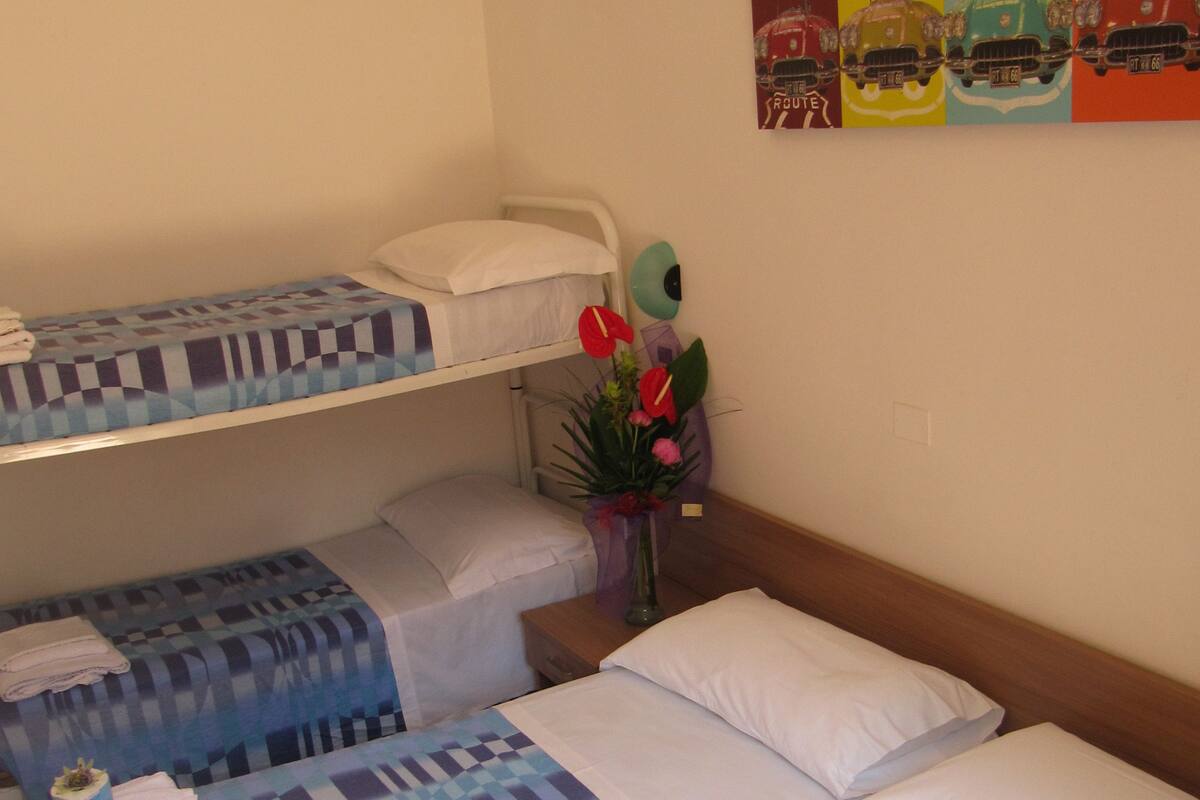 A hotel room with two beds, one with a checkered blue and white bedspread and the other with a plain white bedspread. There is a small table with a vase of flowers and a lamp.