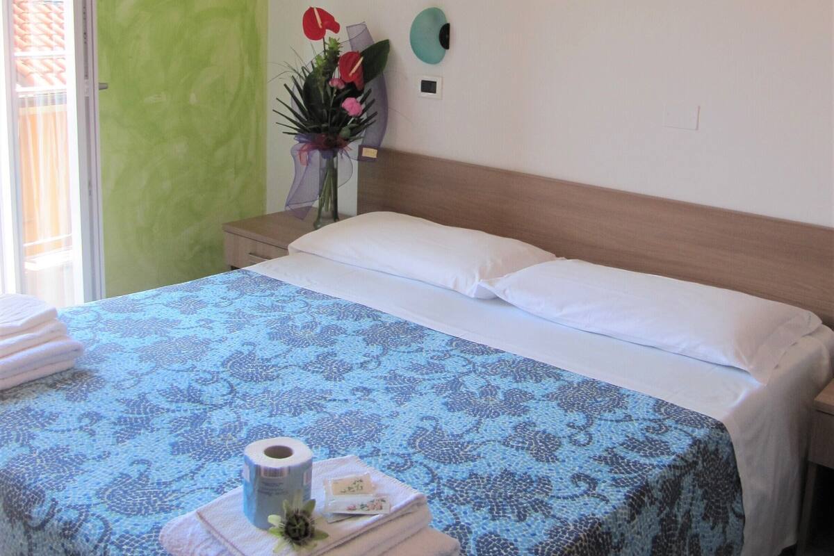 A neatly made bed with a blue patterned bedspread, a white pillow, and a wooden headboard. There is a nightstand with a vase of flowers and a picture frame on the wall.