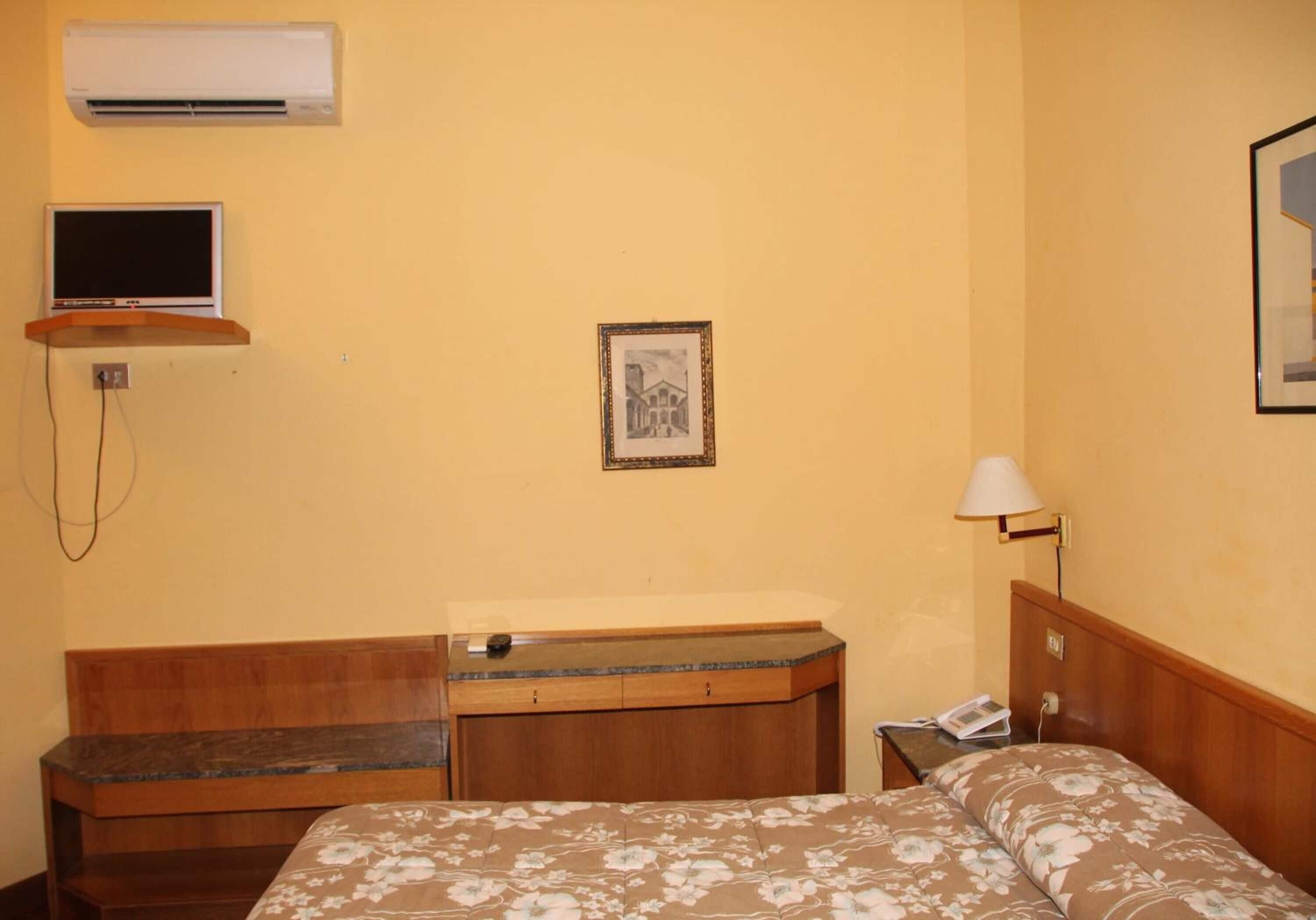 A hotel room with a bed, a wooden bench, a desk, an air conditioner, a television, a framed picture, and a bedside lamp.