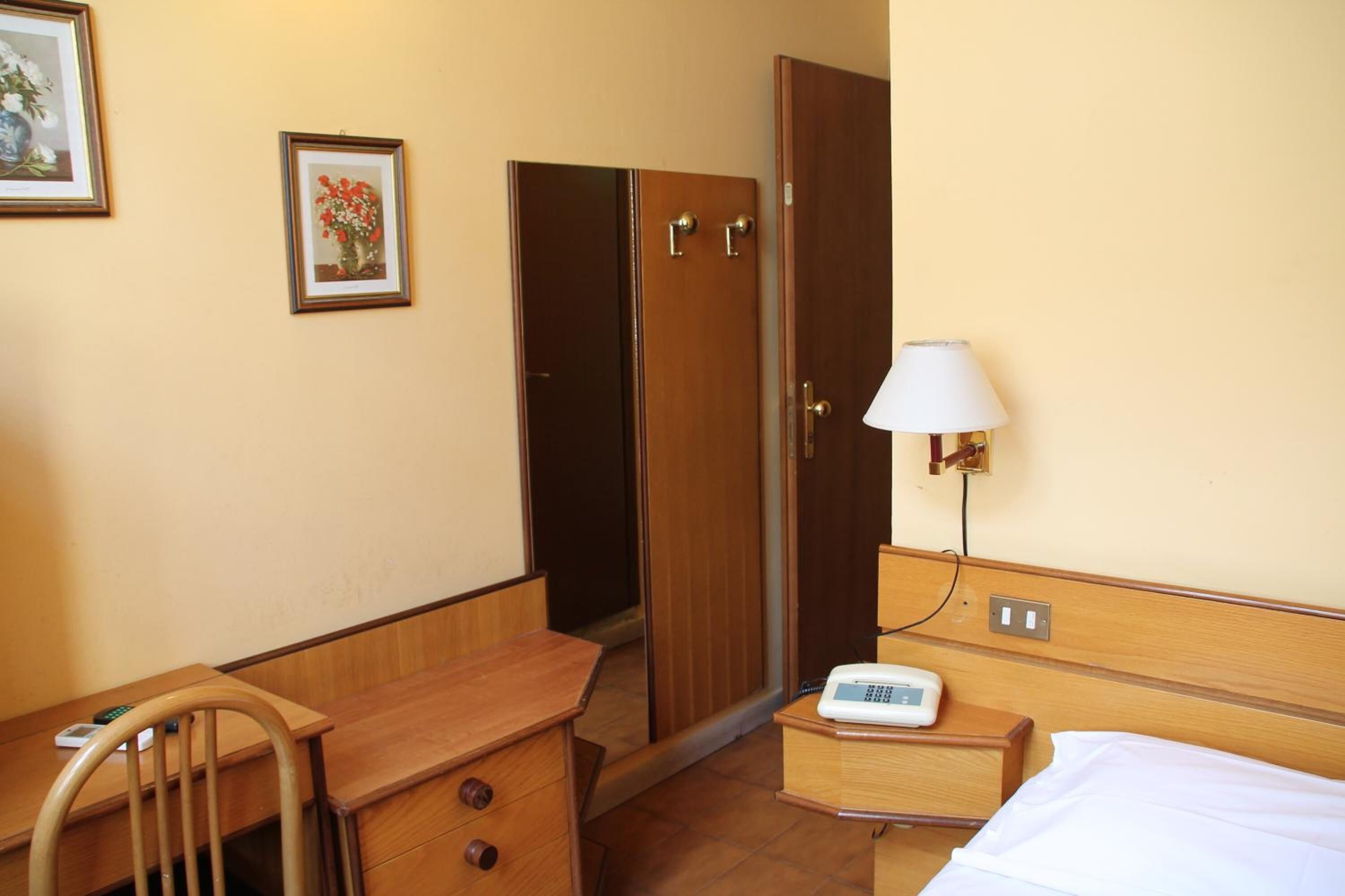 Ein Hotelzimmer mit Holzschreibtisch, einem Sessel, einem Nachttisch mit Lampe, einem Bett und einem Kleiderschrank.