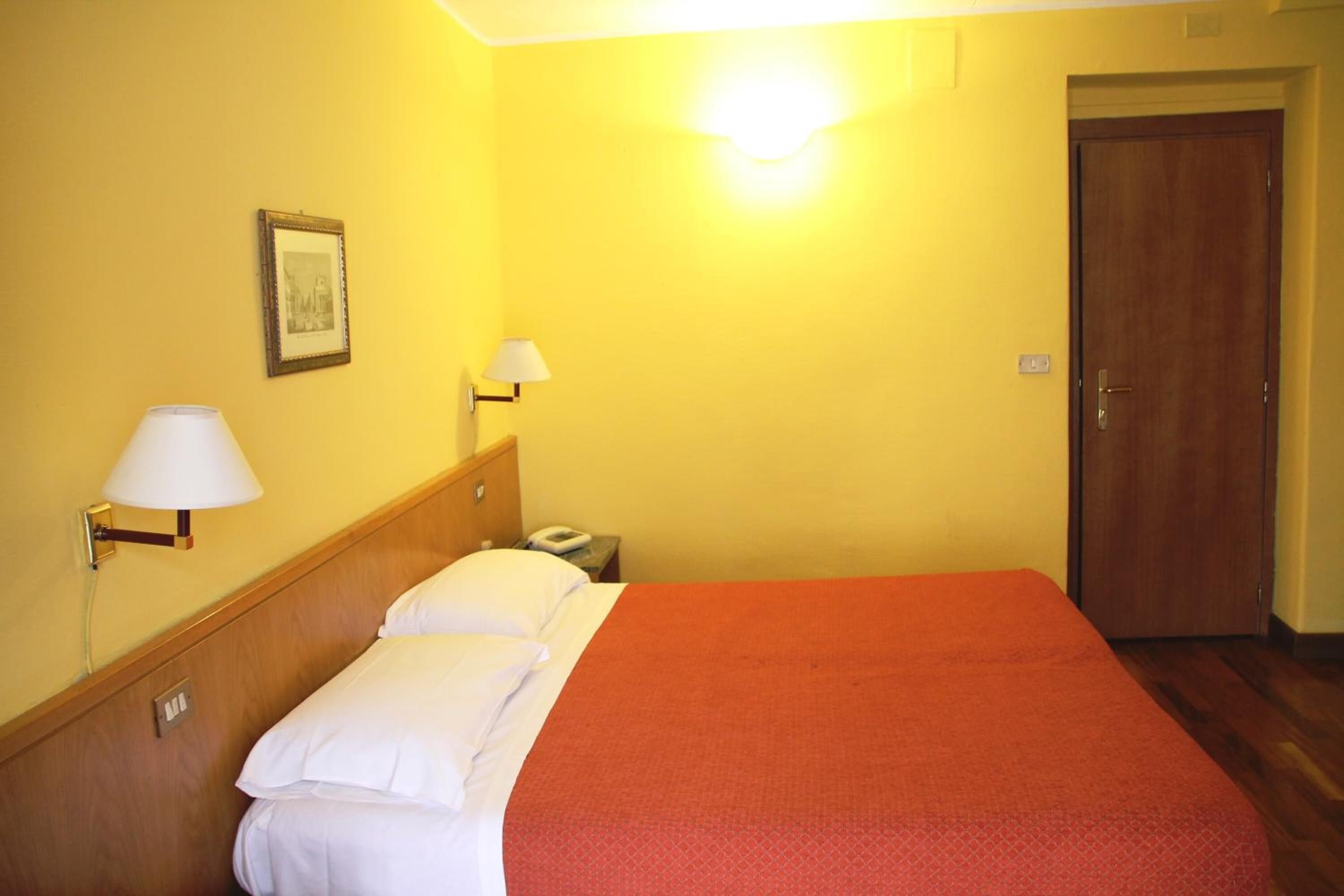 Una habitación de hotel con una cama, dos lámparas de noche, una mesita de noche, un cuadro en la pared y una puerta.