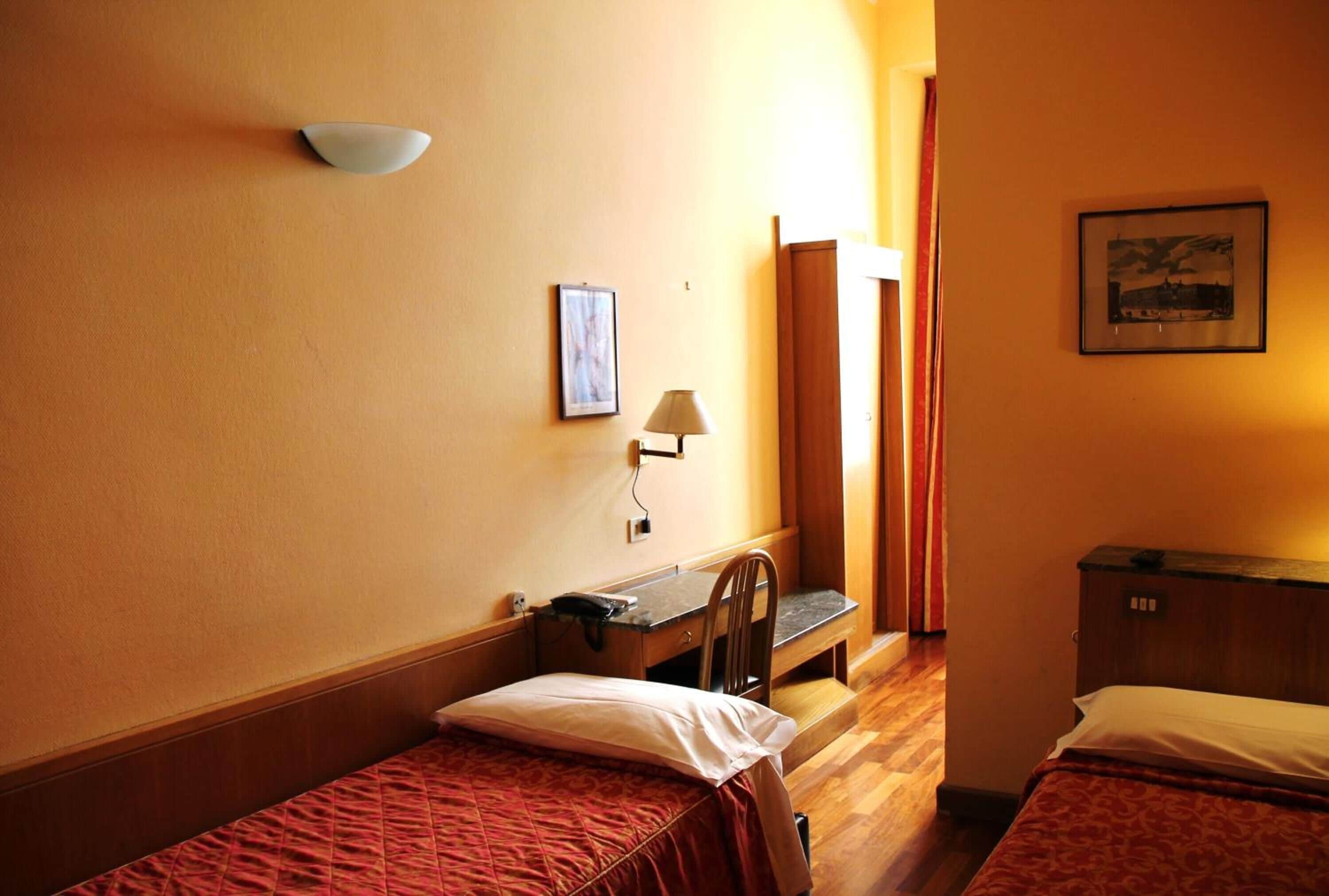 Ein Hotelzimmer mit zwei Einzelbetten, einem Schreibtisch mit Stuhl, einer Lampe und einem Bild an der Wand.