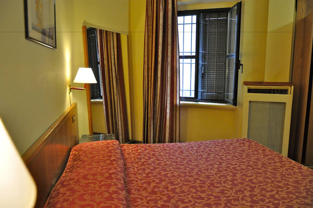 A hotel room with a bed, a nightstand, a lamp, a window with curtains, and a wall-mounted picture.
