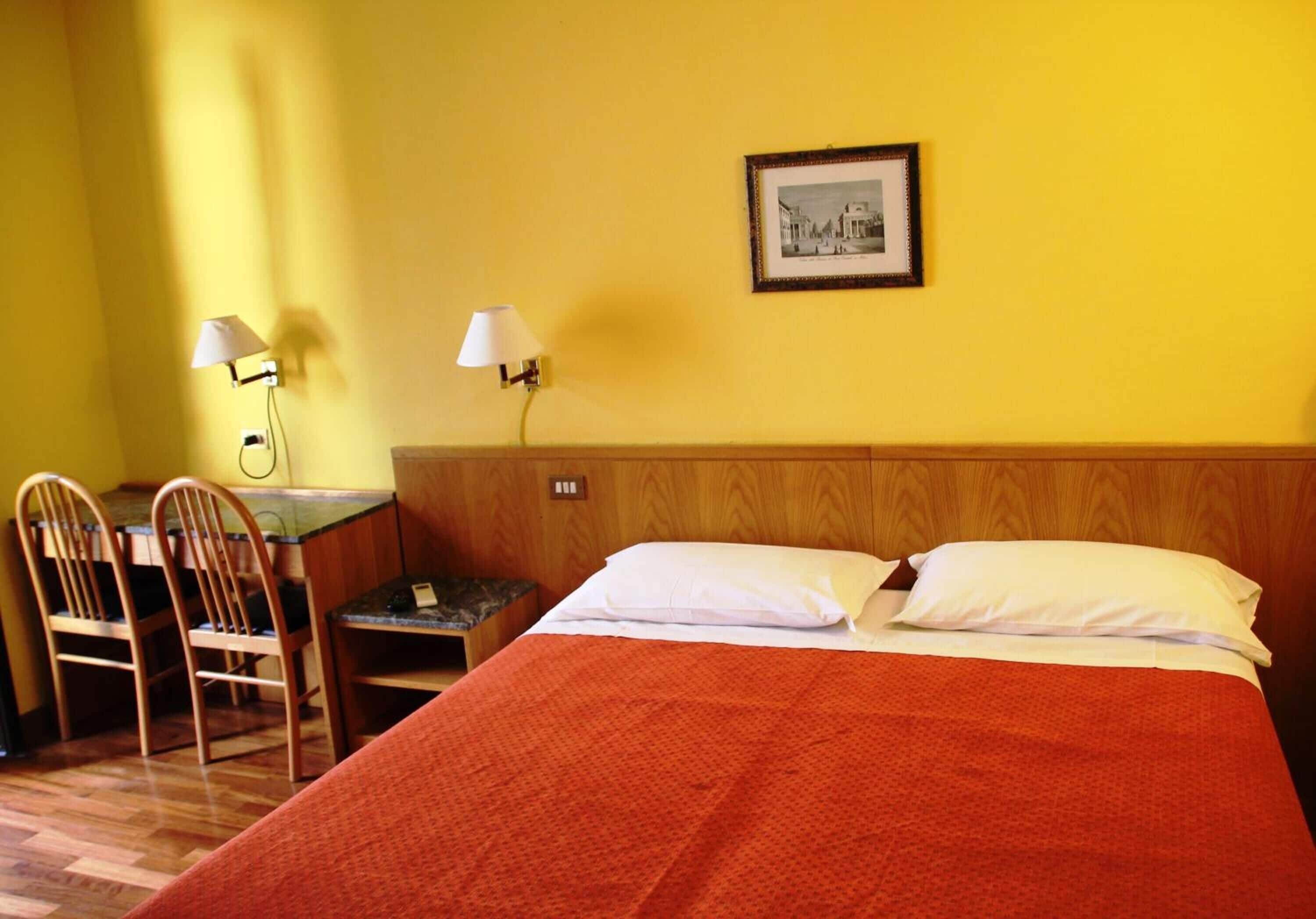 A hotel room with a wooden headboard, a bed with white pillows, two chairs, a small table, and two wall-mounted lamps.