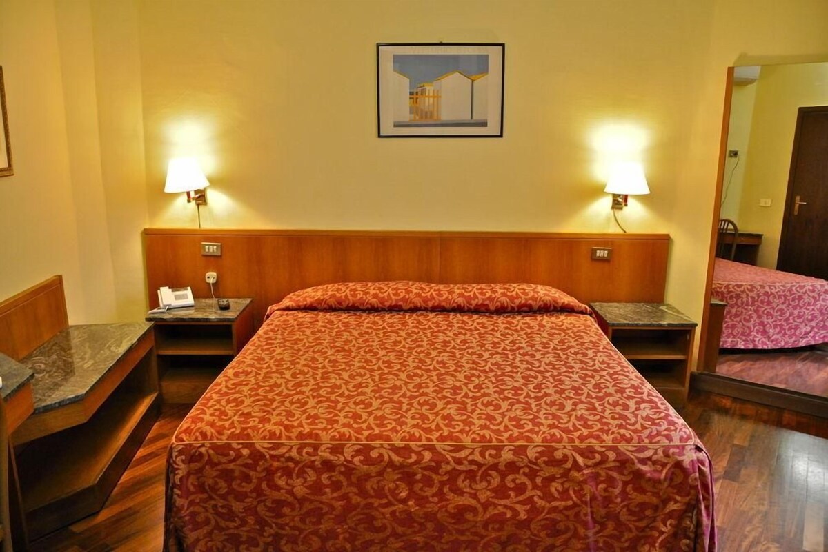 Un cuarto de hotel con una cama grande, dos mesitas de noche, una lámpara y un espejo. En la pared, un cuadro enmarcado.