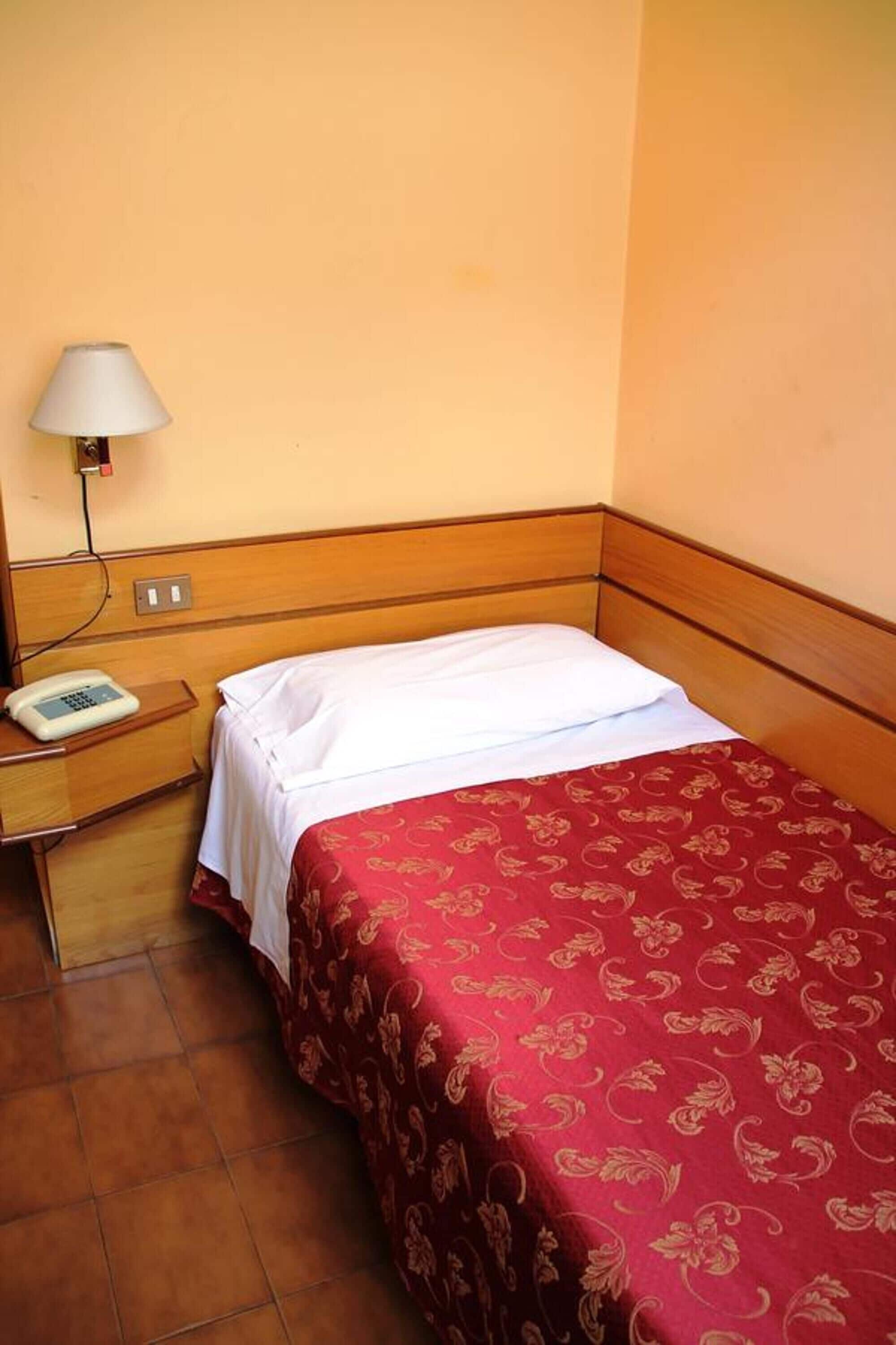 A hotel room with a wooden headboard, a single bed with a red patterned bedspread, a white pillow, a bedside table with a lamp, and a telephone.