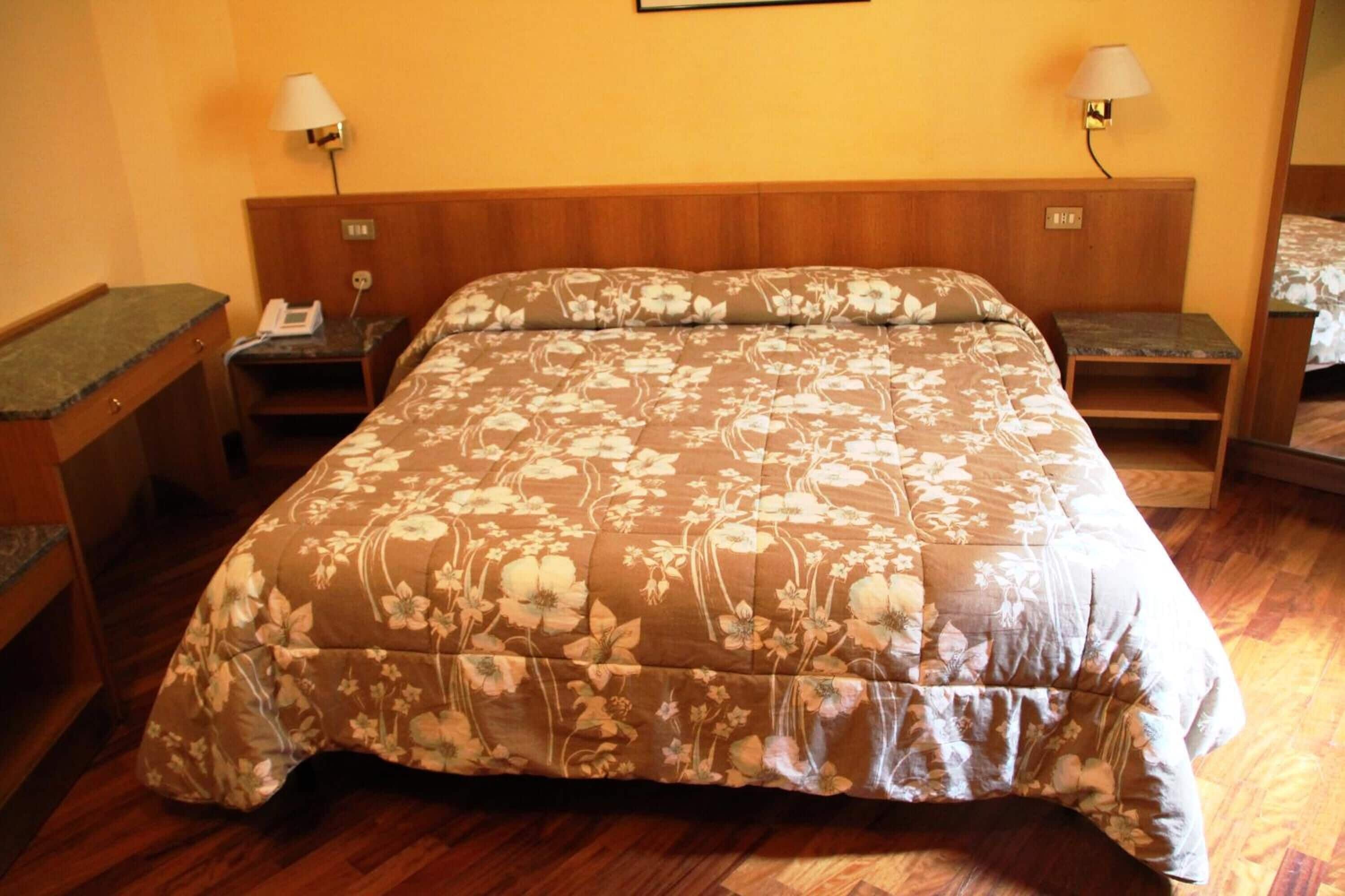A hotel room with a large bed, wooden headboard, two bedside tables with lamps, a telephone, and a small desk with a lamp.