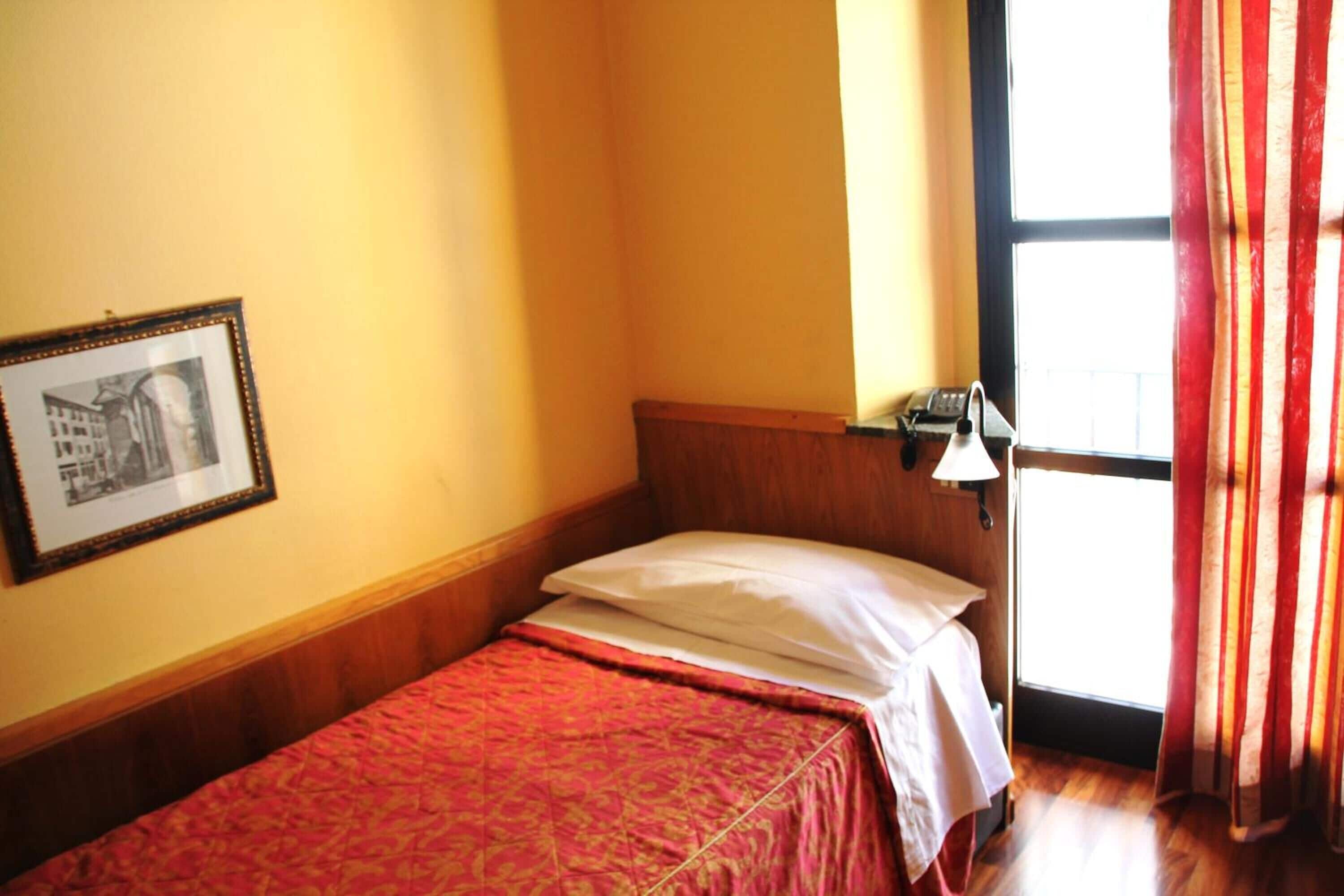 A small, single-bed room with a wooden headboard, a framed picture on the wall, a bedside lamp, and a window with curtains.