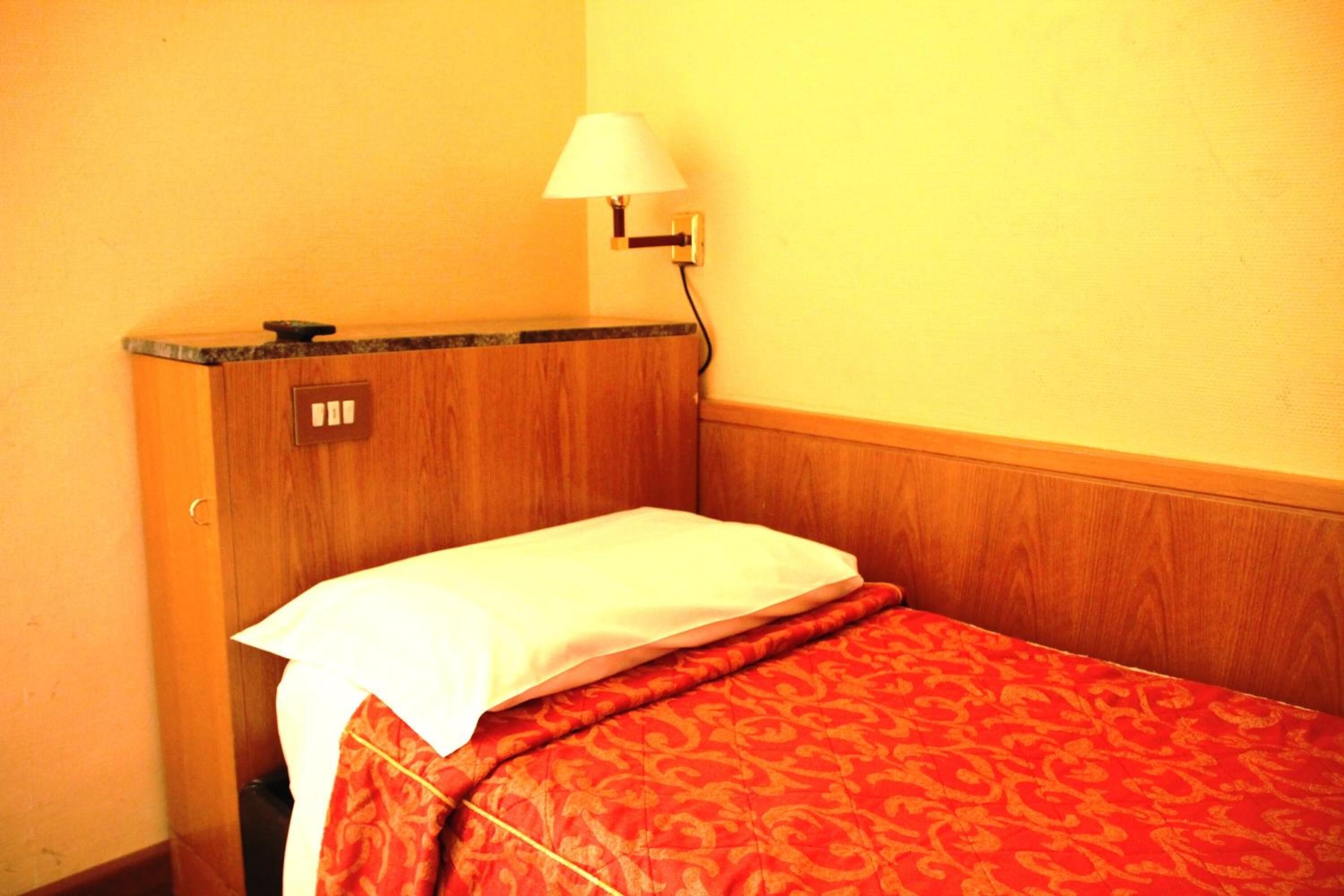 A small, single-bed room with a wooden headboard, a bedside lamp, and a patterned bedspread.