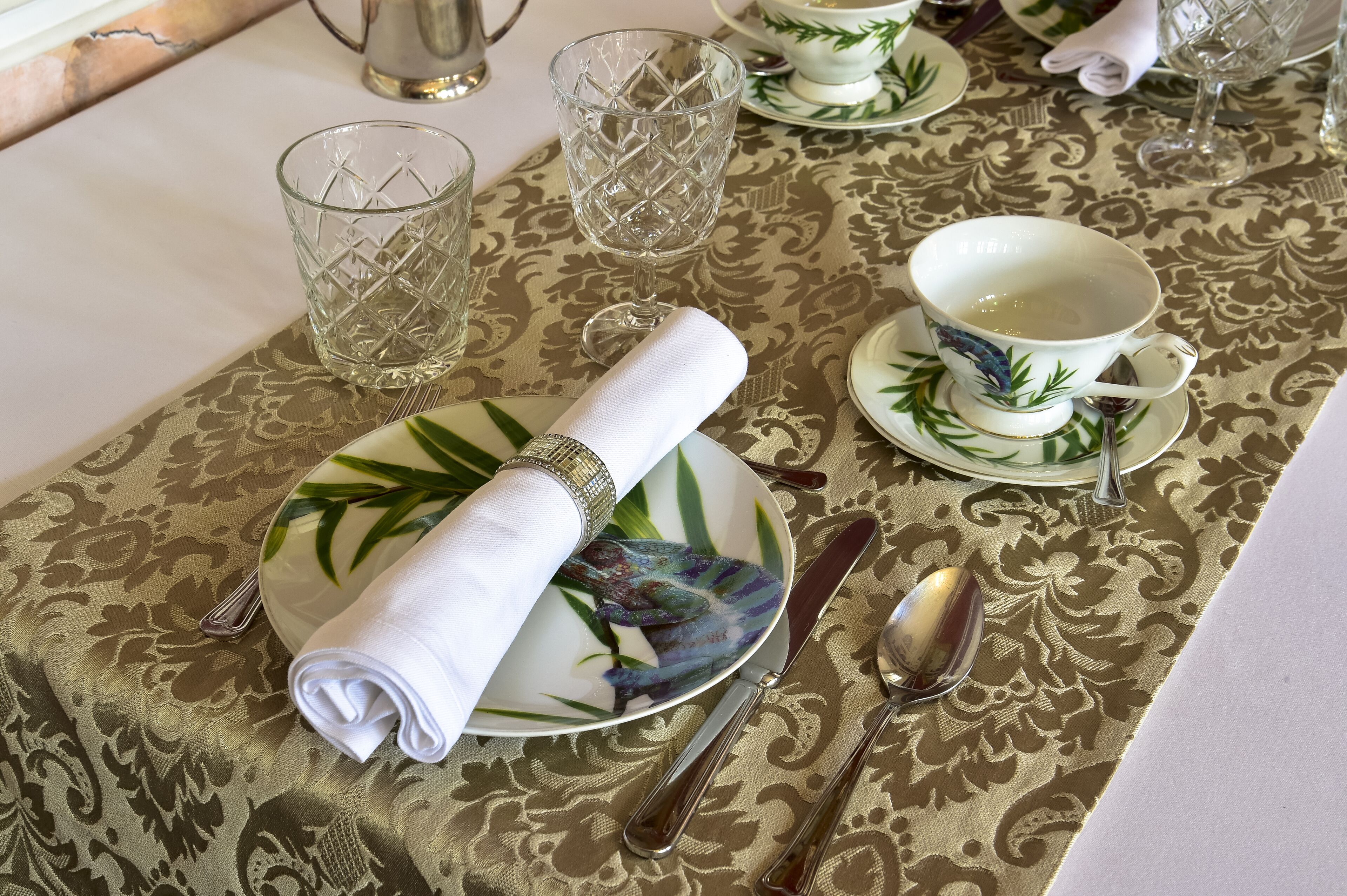 Une table dressée avec une serviette blanche pliée, une assiette ornée d’un motif de feuille verte, une tasse et des couverts.