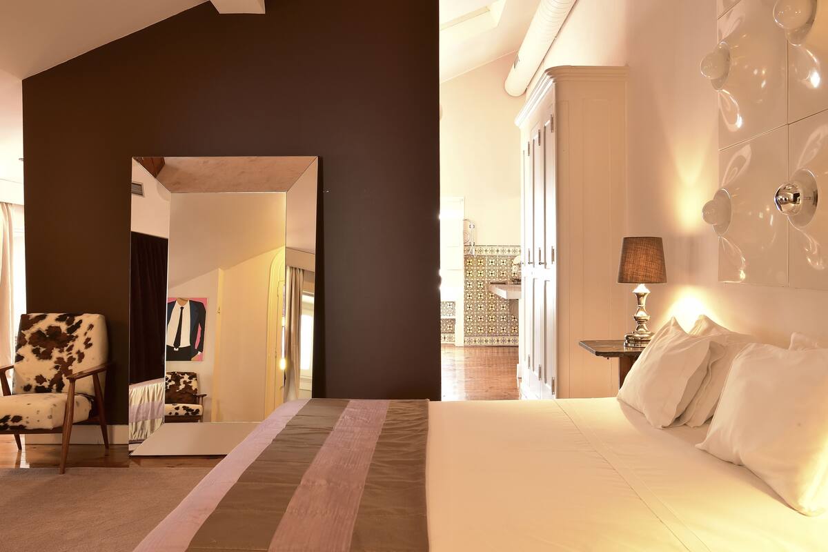 A modern hotel room with a large bed, a chair, and a mirror reflecting the room.