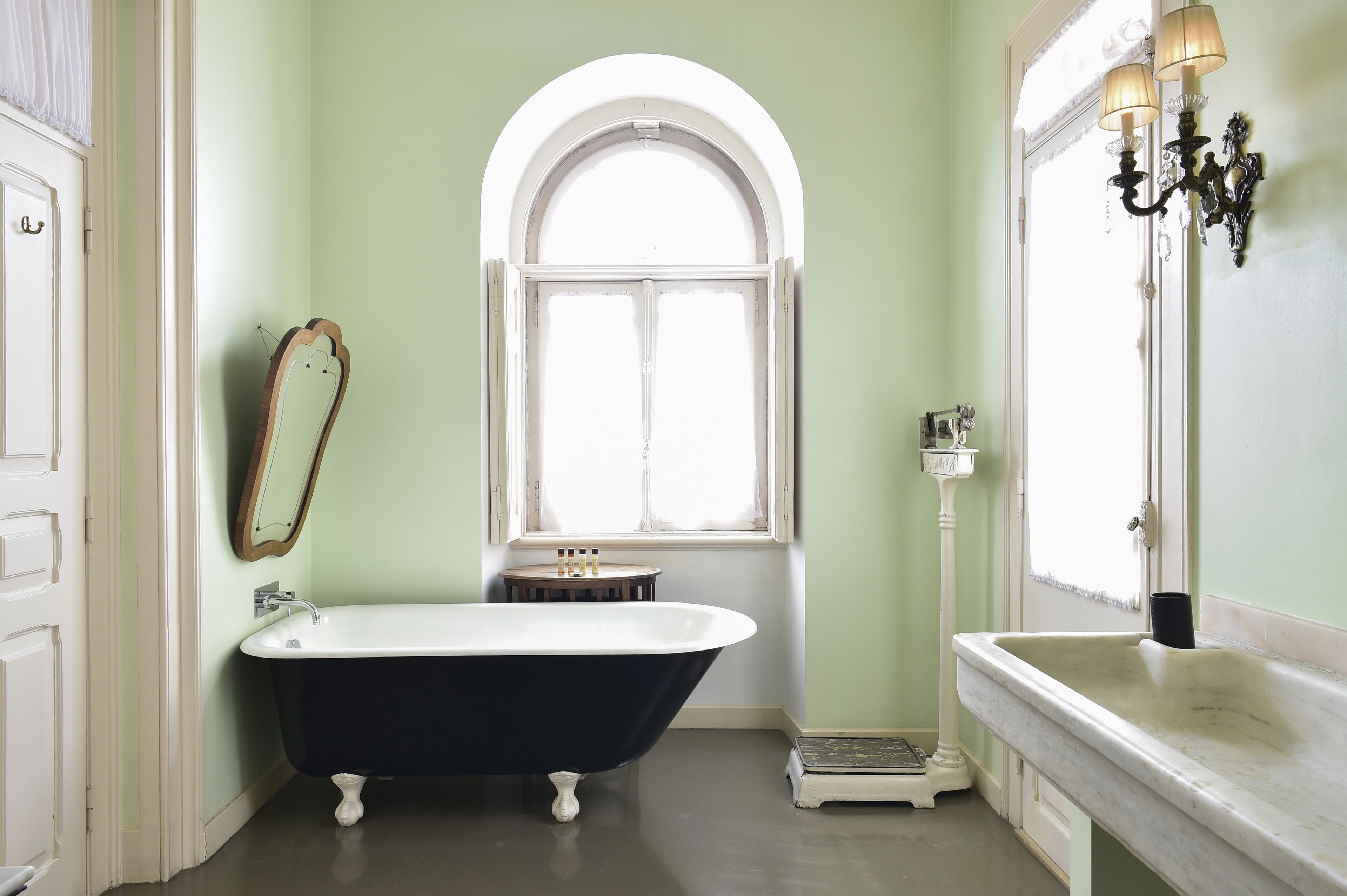 A bathroom with a freestanding bathtub, a window with an arched top, a vanity with a mirror, and a wall-mounted light fixture.