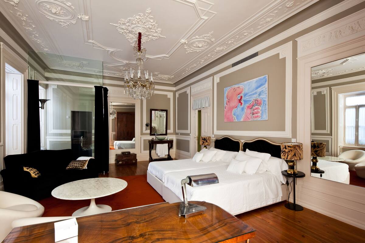 A luxurious hotel room with a large bed, a sitting area with a round table, a chandelier, and a painting on the wall.