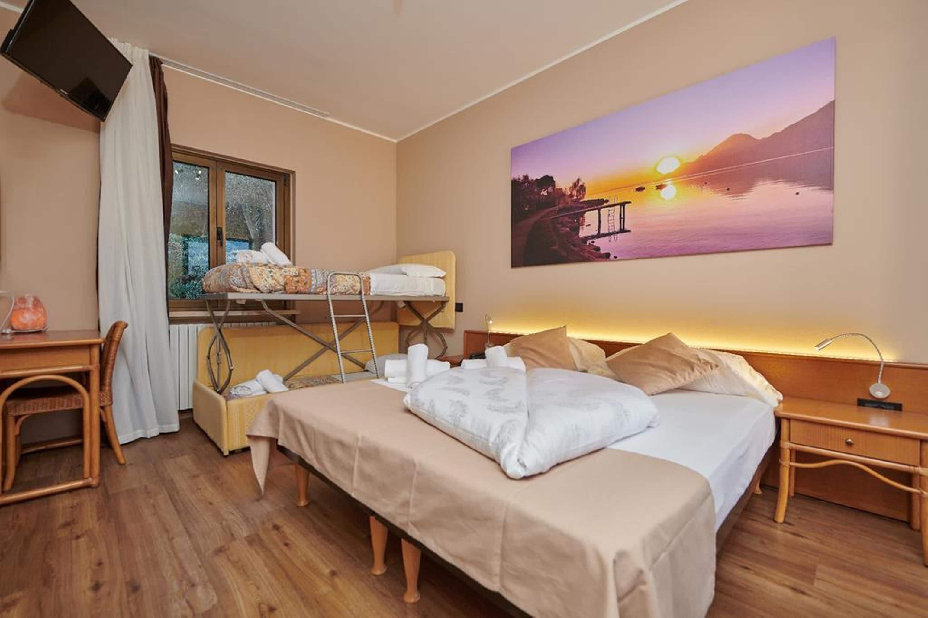 A hotel room with a bed, a desk, a chair, a window with curtains, and a painting of a sunset.