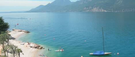 A coastal beach with clear waters, boats, and mountains in the background.