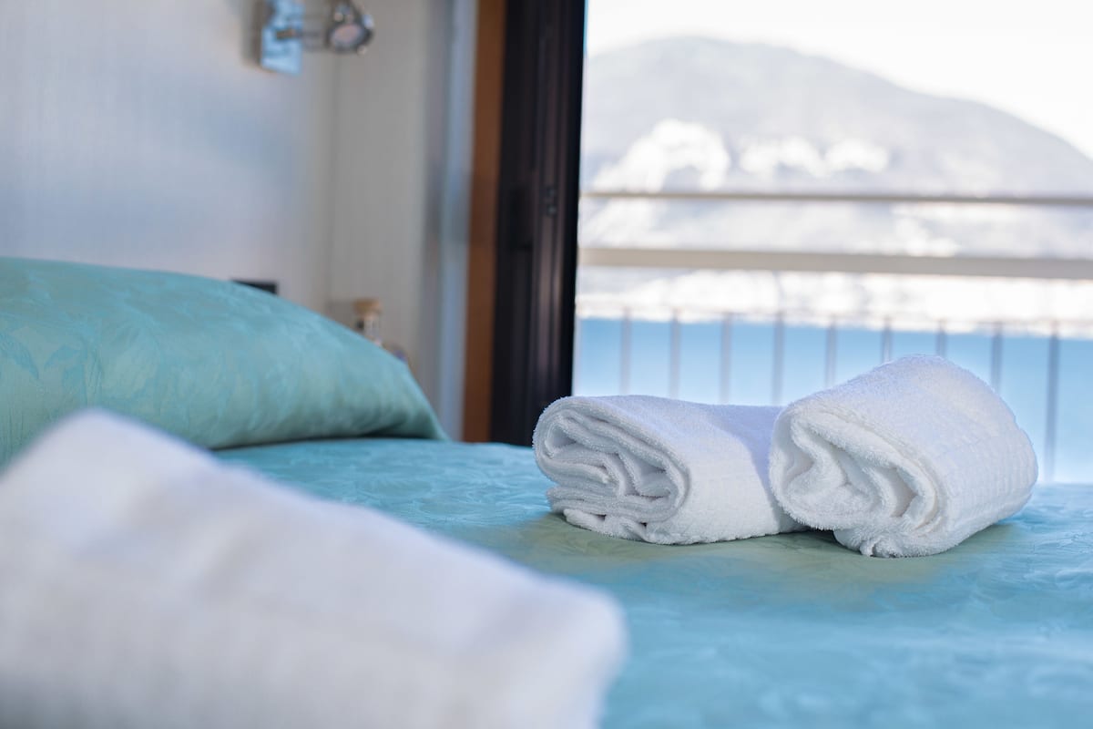 Ein ordentlich bezogenes Bett mit einem gefalteten weißen Handtuch und einem blauen Kopfkissen, mit Blick auf einen verschneiten Berg und einer Geländer im Hintergrund.