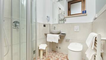 Un baño con cabina de ducha de vidrio, lavamanos con espejo, inodoro y un pequeño taburete.