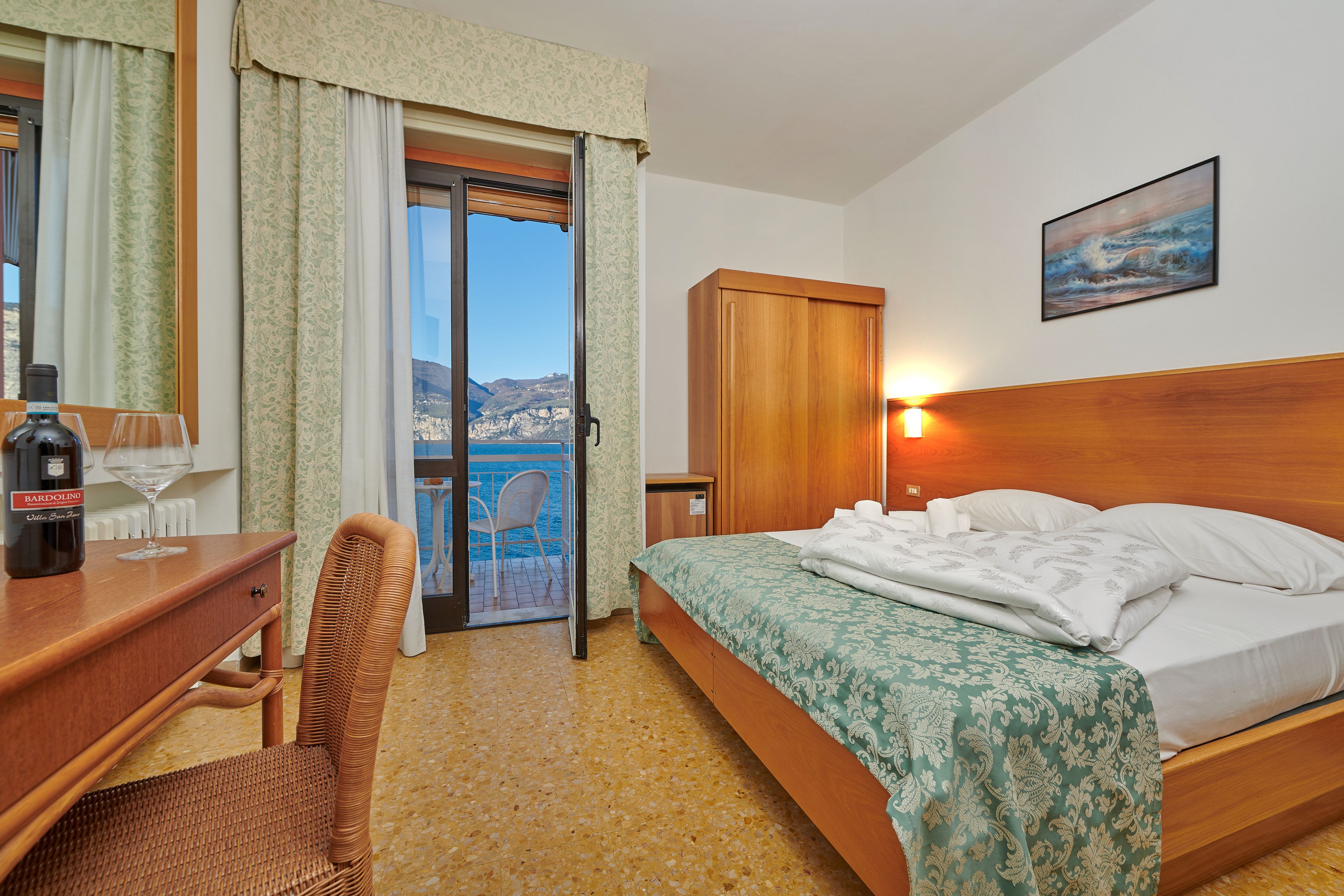 Habitación de hotel con cama, escritorio con botella de vino, una silla, un cuadro en la pared y vista al mar desde la ventana.