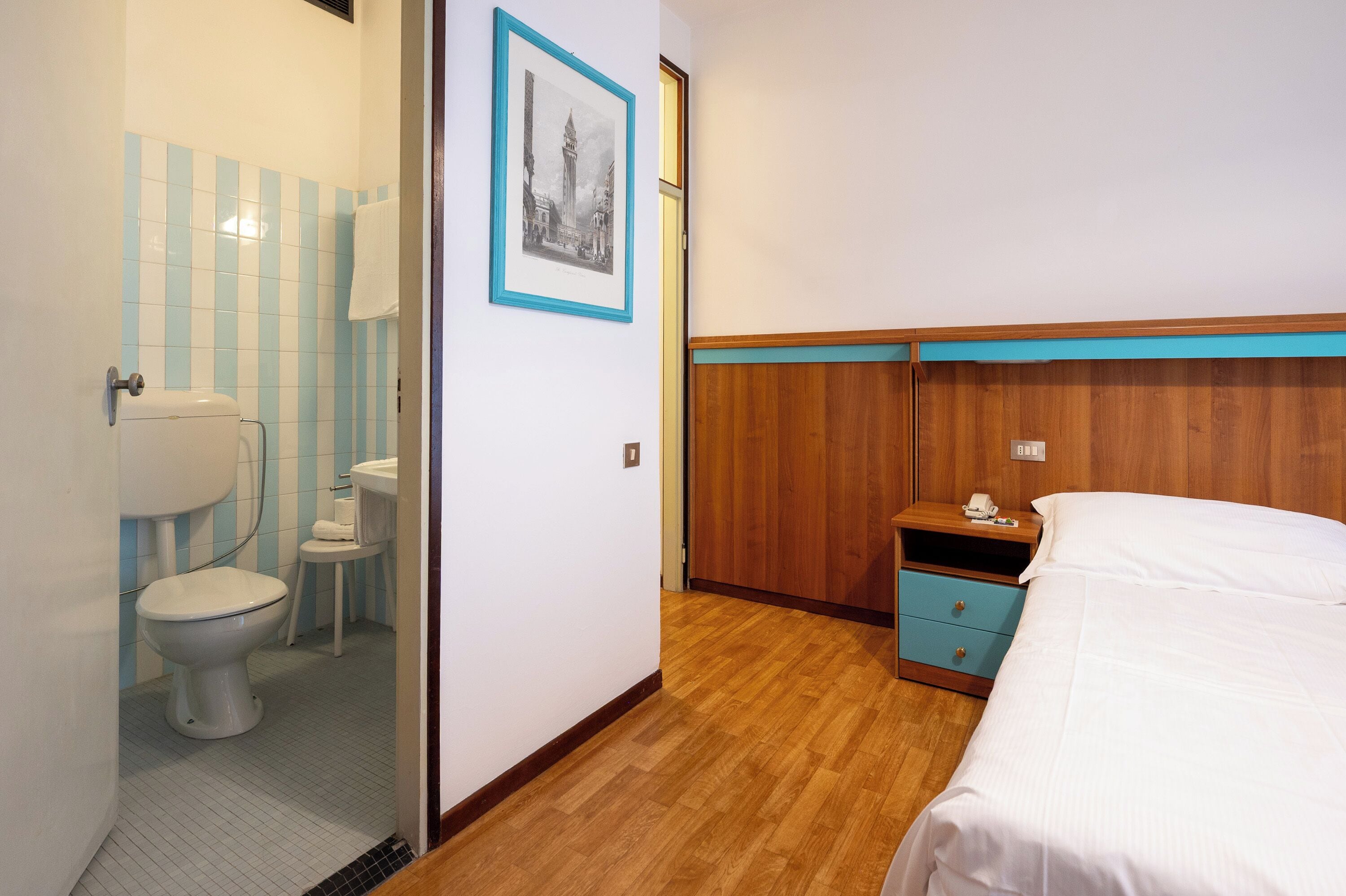 A hotel room with a wooden headboard, a bed, a nightstand with a phone, a small table with a lamp, and a bathroom with a toilet and a sink.