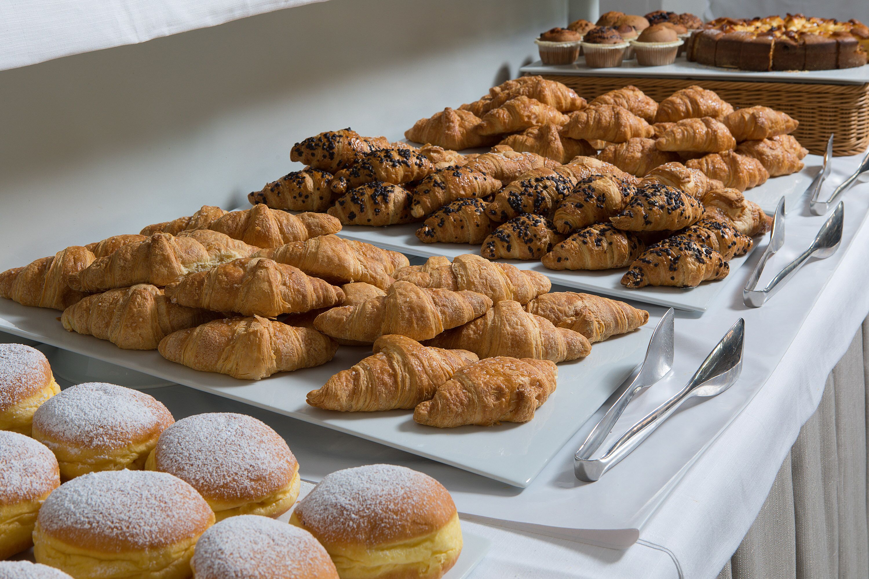 A buffet table with various pastries and bread, including croissants and muffins, arranged neatly on white plates.