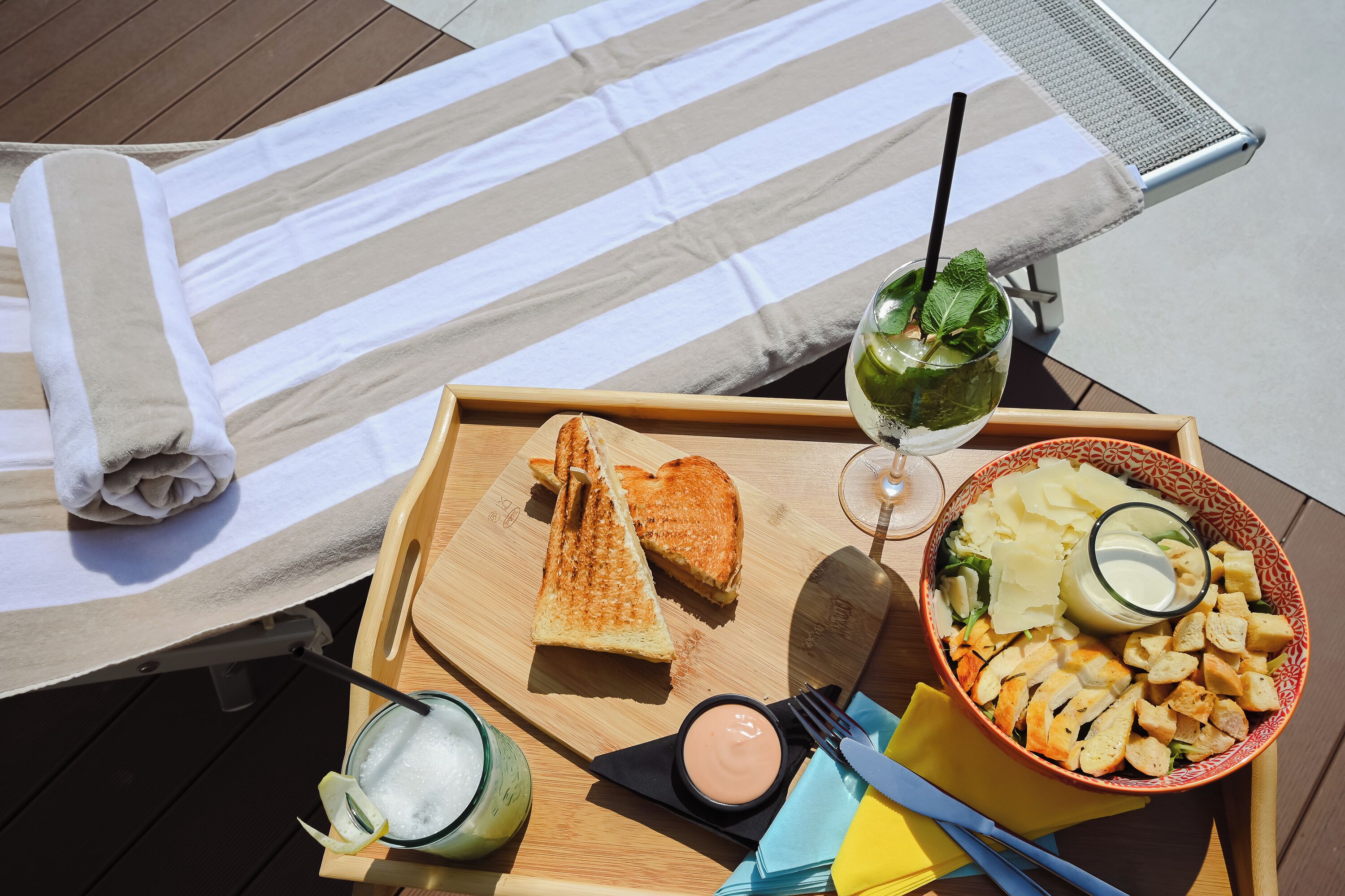 Un plateau en bois avec un sandwich au fromage grillé, un saladier rempli de salade et une boisson, le tout posé sur une terrasse.