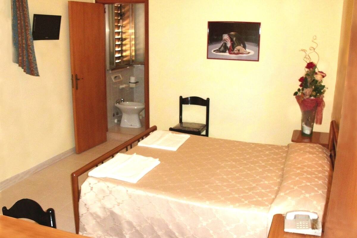 Habitación de hotel con una cama, una silla, una mesita de noche con un teléfono, un cuadro en la pared y un baño visible a través de una puerta abierta.