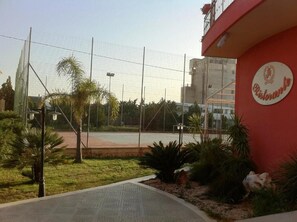 Un terrain de sport entouré d’une clôture, avec des palmiers et un bâtiment sur lequel on peut lire l’inscription « Ristorante ».