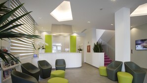 A modern reception area with a curved desk, grey and green chairs, and a staircase with pink steps. - Hotel Gardenia (Jesolo)