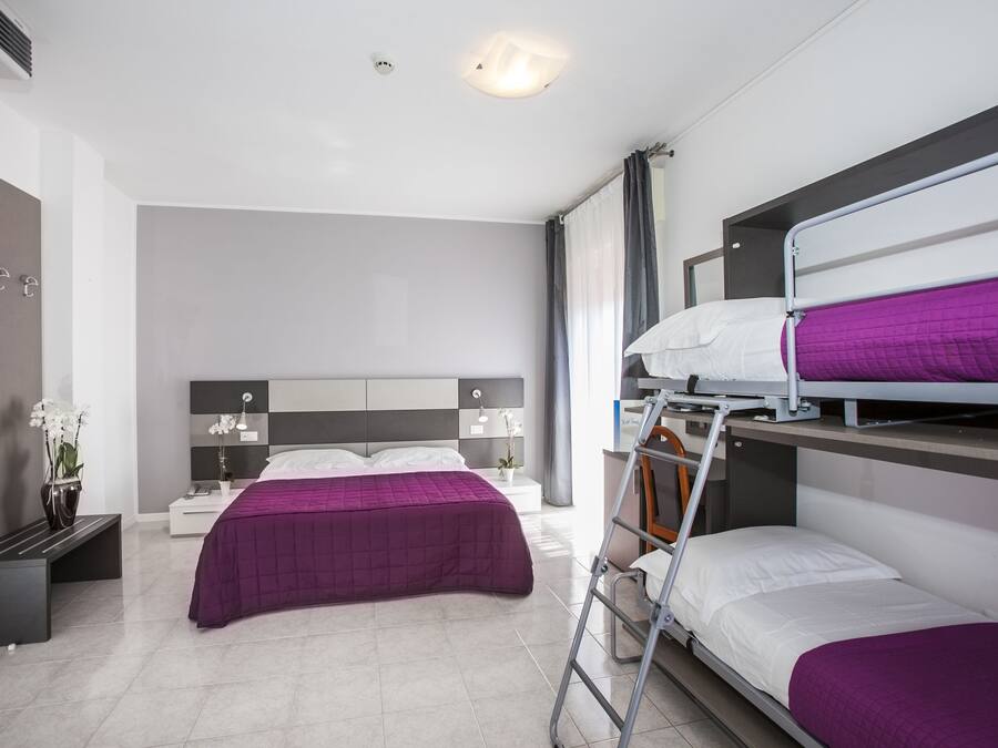 A hotel room with a bunk bed, a single bed, a nightstand, a lamp, a vase with flowers, and a bench.