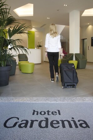 Un hall d’hôtel avec un comptoir d’accueil, des sièges verts et un paillasson sur lequel est inscrit « Hôtel Gardenia ».
