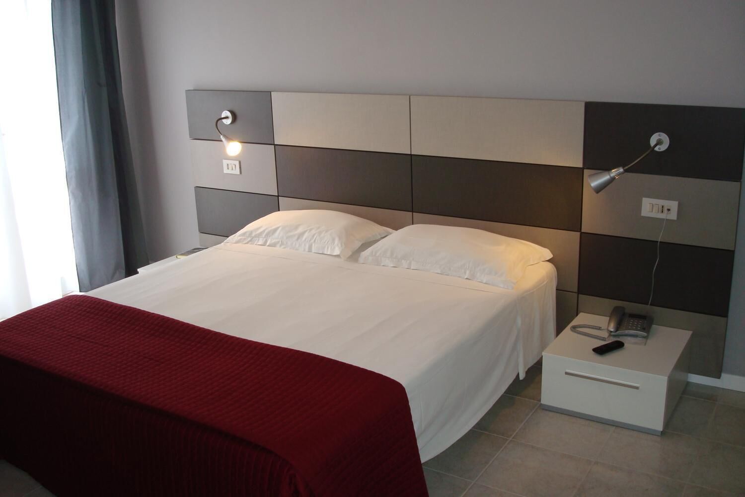Un lit bien fait, avec du linge de lit blanc, un plaid rouge, une table de chevet avec un téléphone et une télécommande, et une lampe fixée au mur.