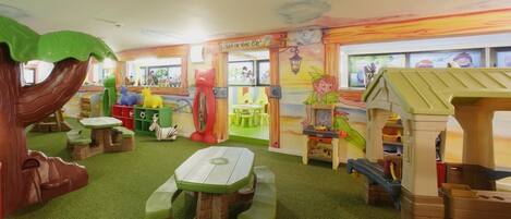 Zona de juegos infantiles con una casita de madera, mesas y una gran estructura arbórea.