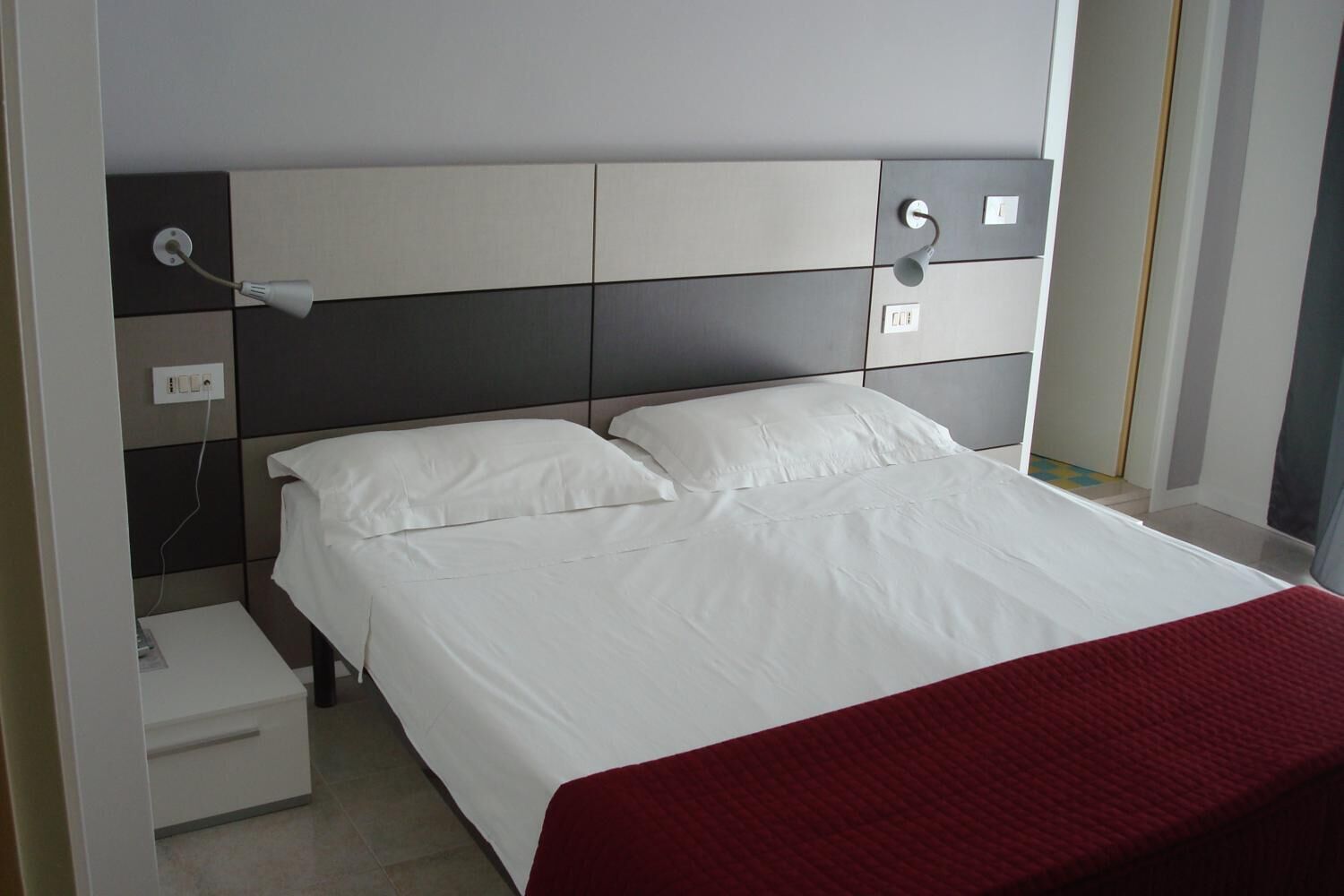 Un lit bien fait, avec du linge de lit blanc et une couverture rouge, une table de chevet avec une lampe, et un luminaire fixé au mur.