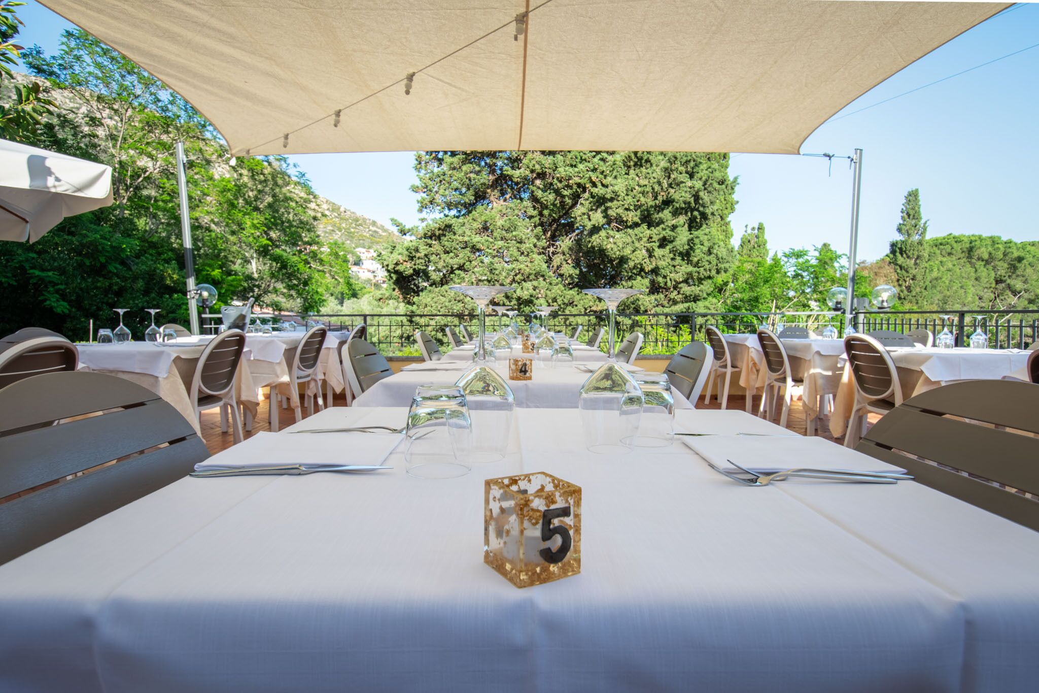 A table set for a meal with a number 5 sign, surrounded by chairs and tables under a canopy, with trees in the background.