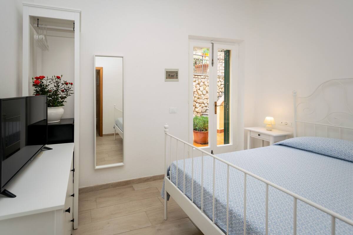 A bedroom with a bed, bedside table, lamp, and a TV on a desk.