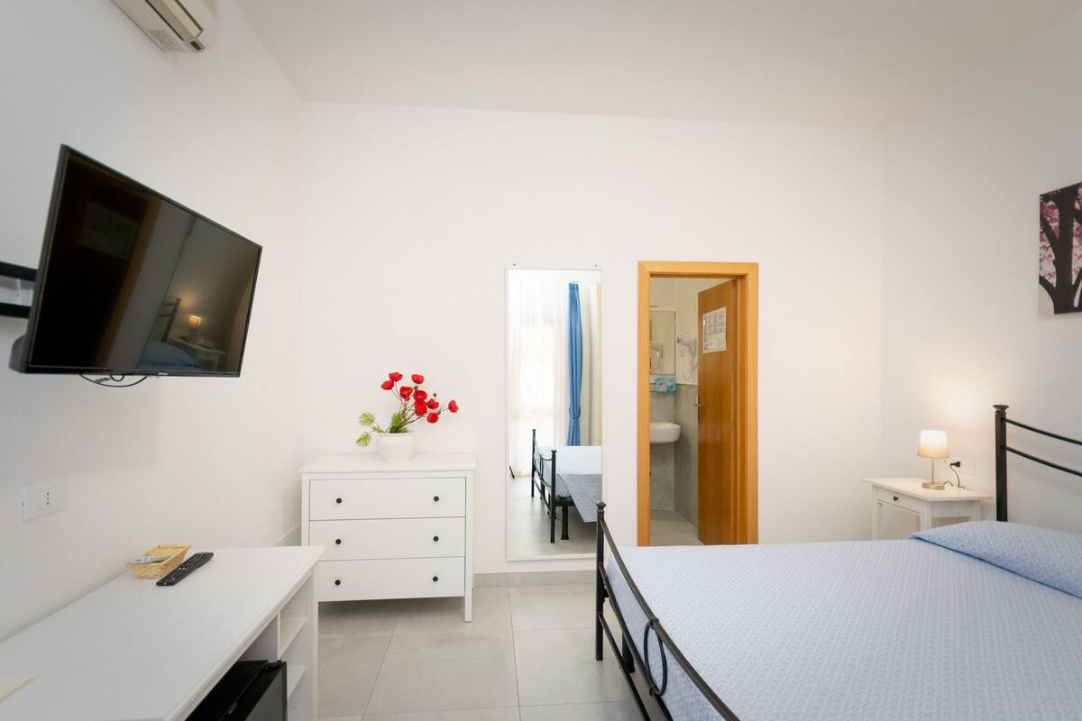 A bedroom with a bed, a desk, a dresser, a TV, and a bathroom visible through an open door.