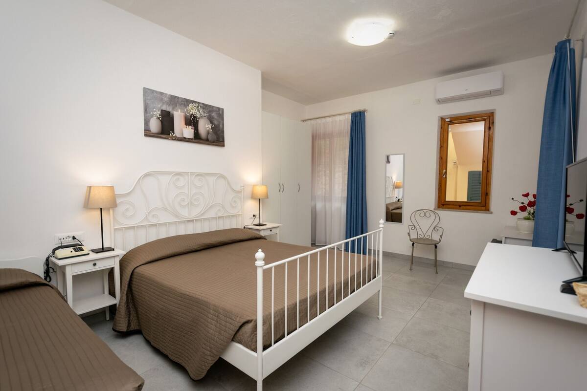 A bedroom with a bed, bedside tables, a desk, a chair, a window with curtains, and a wall-mounted air conditioner.